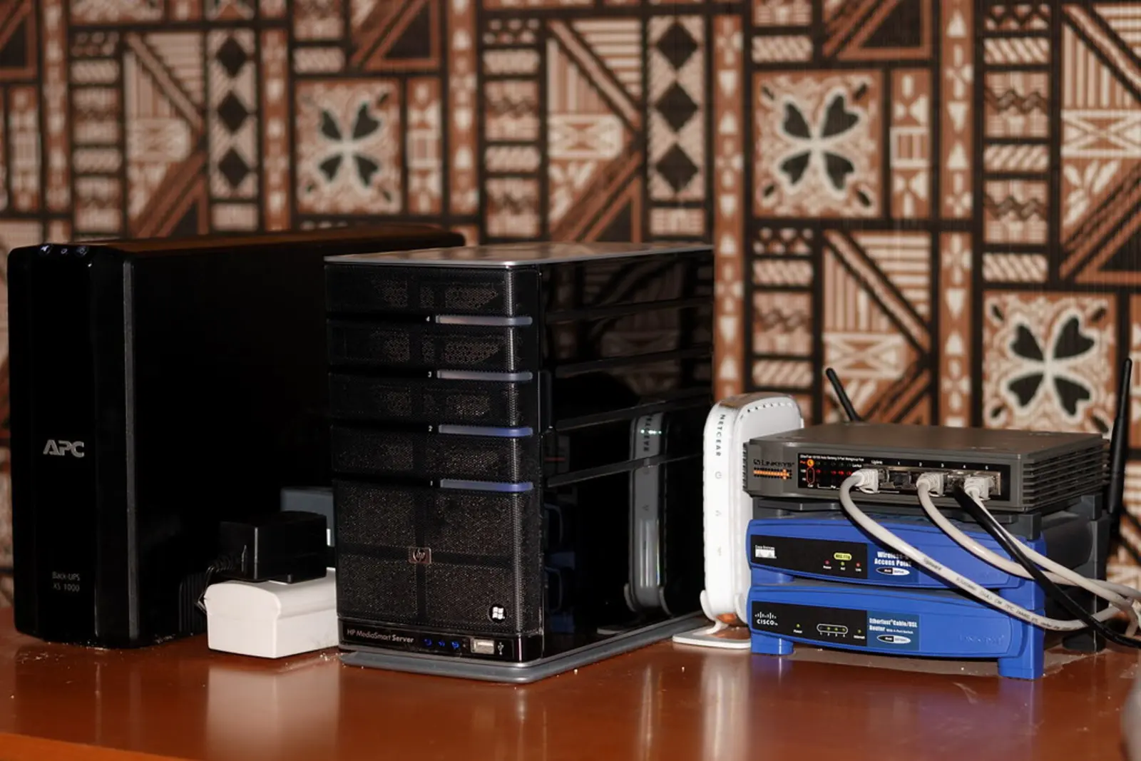 A black APC uninterruptible power supply stands next to a server tower and network switches on a wooden desk.