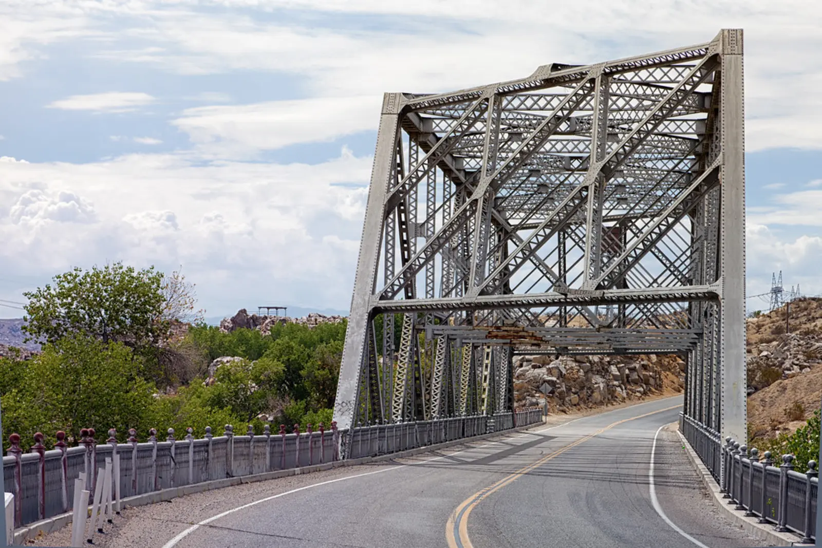 A gray steel truss bridge featuring a complex lattice structure spans a paved two-lane road under a bright sky with scattered clouds.