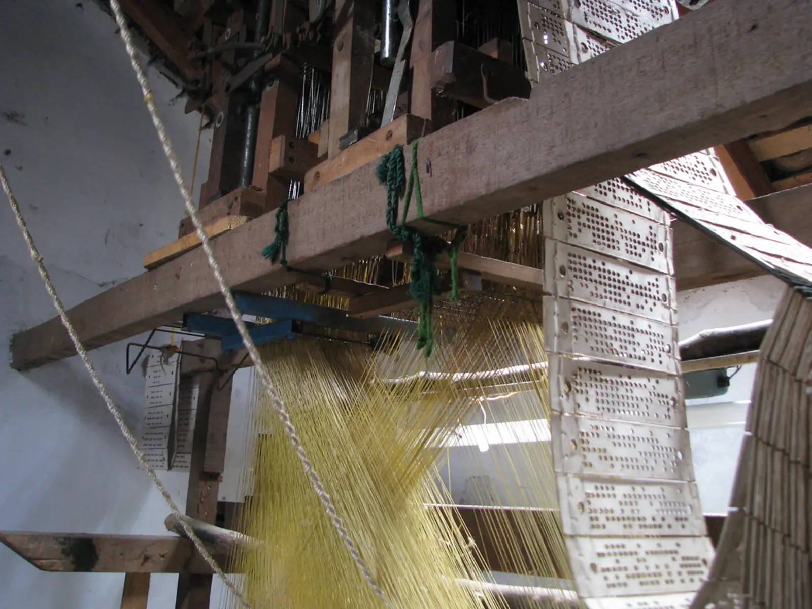 A wooden hand-loom with yellow threads and a row of perforated white cards hanging on the right side. The photographer titled it “India - Sights & Culture - Complicated hand-loom for silk weaving”.