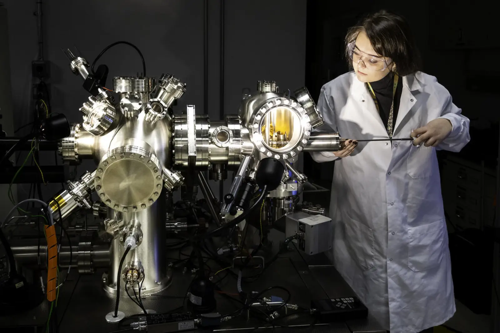 A researcher in a white lab coat and safety glasses inserts a long metal tool into a complex stainless-steel vacuum-chamber apparatus fitted with multiple ports and flanges. The photographer titled it “Functional Atomic Force Microscopy Group research lab”.