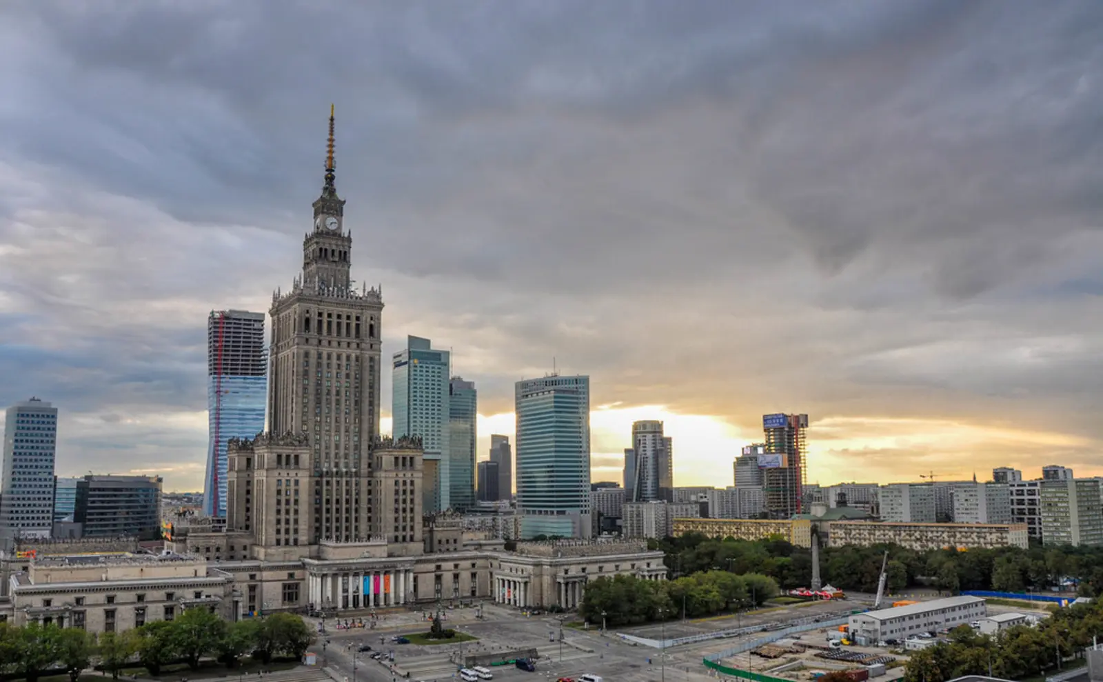 A city skyline featuring a tall, ornate, multi-tiered skyscraper with a spire, surrounded by modern glass high-rise buildings under a cloudy sky. The photographer titled it “Palace of Culture and Science”.
