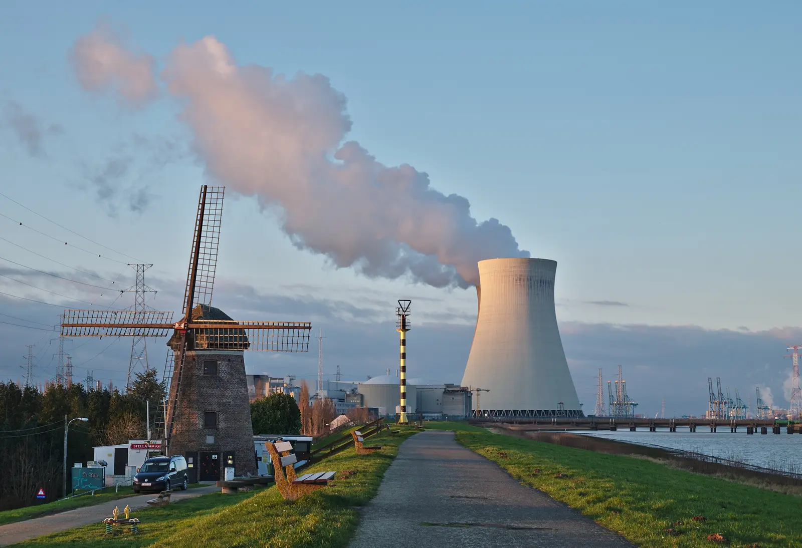 A traditional windmill stands in the foreground while a large nuclear power plant cooling tower emits a thick plume of steam in the background. The photographer titled it “De Molen (windmill) and the nuclear power plant cooling tower in Doel, Belgium (DSCF3859)”.