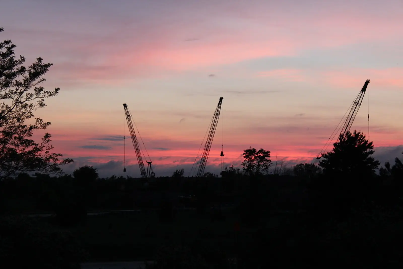 Silhouetted construction cranes stand against a pink and orange sunset sky above a dark landscape of trees. The photographer titled it “End of Shift”.