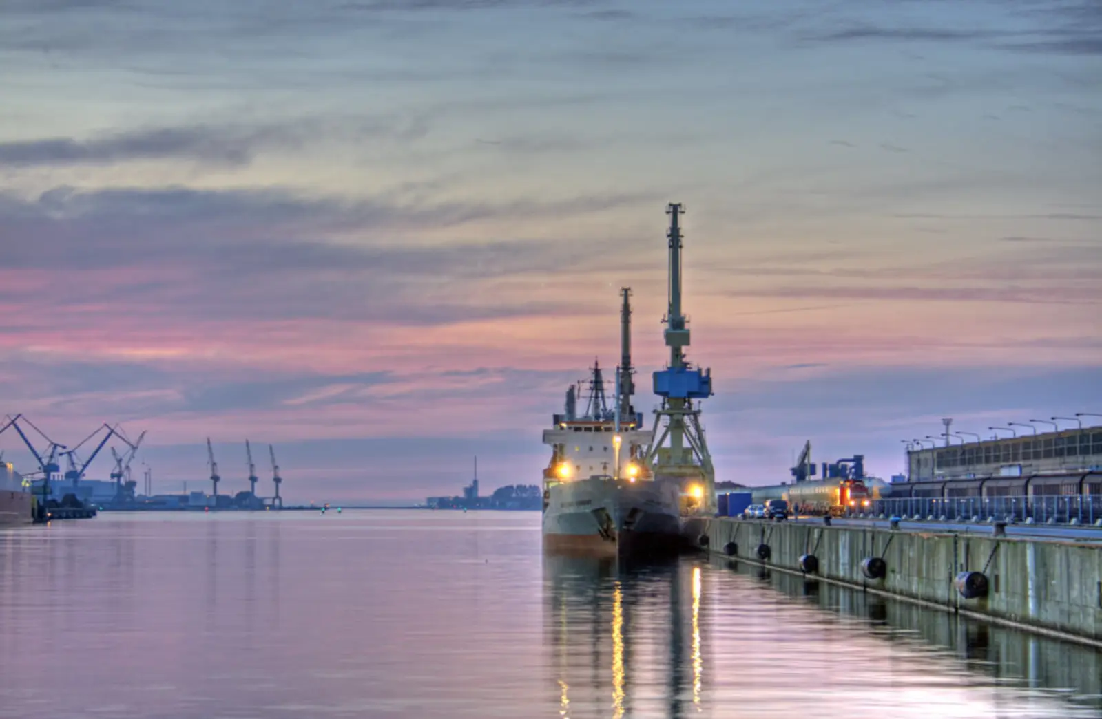 A large cargo ship is docked at a concrete pier in a harbor, with several cranes visible in the background under a pastel twilight sky. The photographer titled it “Waiting for the ferry”.