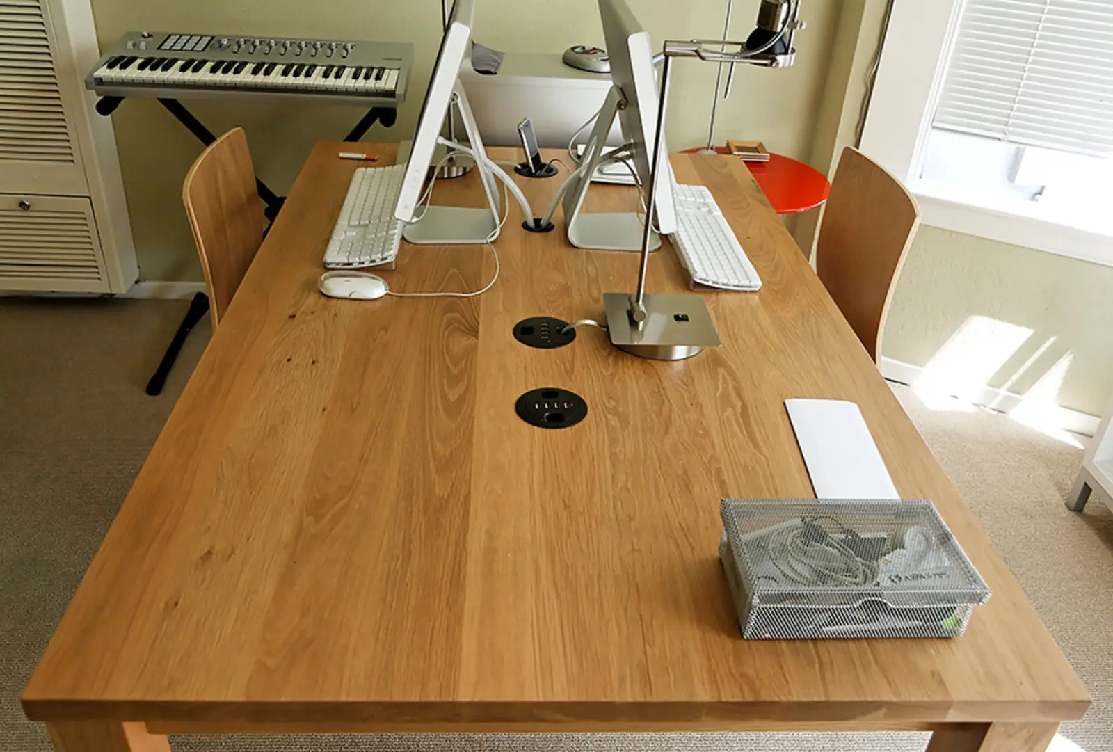 A light wooden table serves as a workspace with two desktop computers, keyboards, a mouse, a metal lamp, and a wire mesh basket, with a musical keyboard on a stand in the background. The photographer titled it “Desk | San Francisco”.