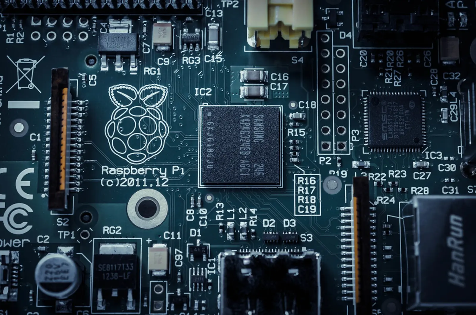 A close-up view of a green circuit board featuring a central black square microchip, gold edge connectors, and various small electronic components. The photographer titled it “Raspberry Pi”.