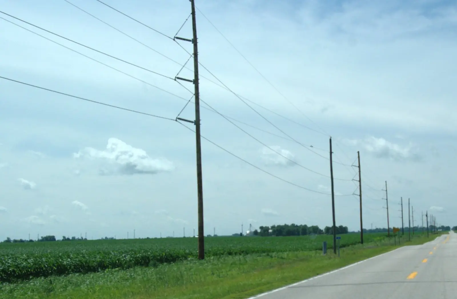 A row of wooden utility poles carrying power lines runs alongside a paved rural road next to a green field under a cloudy sky. The photographer titled it “Wind farm 138kV lines”.