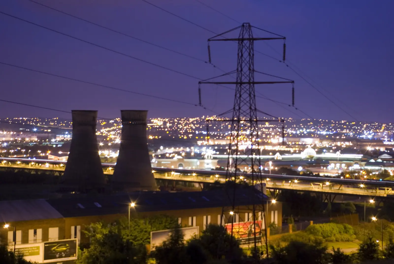 A high-voltage transmission tower stands in the foreground at dusk, with industrial cooling towers and a brightly lit city skyline in the background. The photographer titled it “Power”.