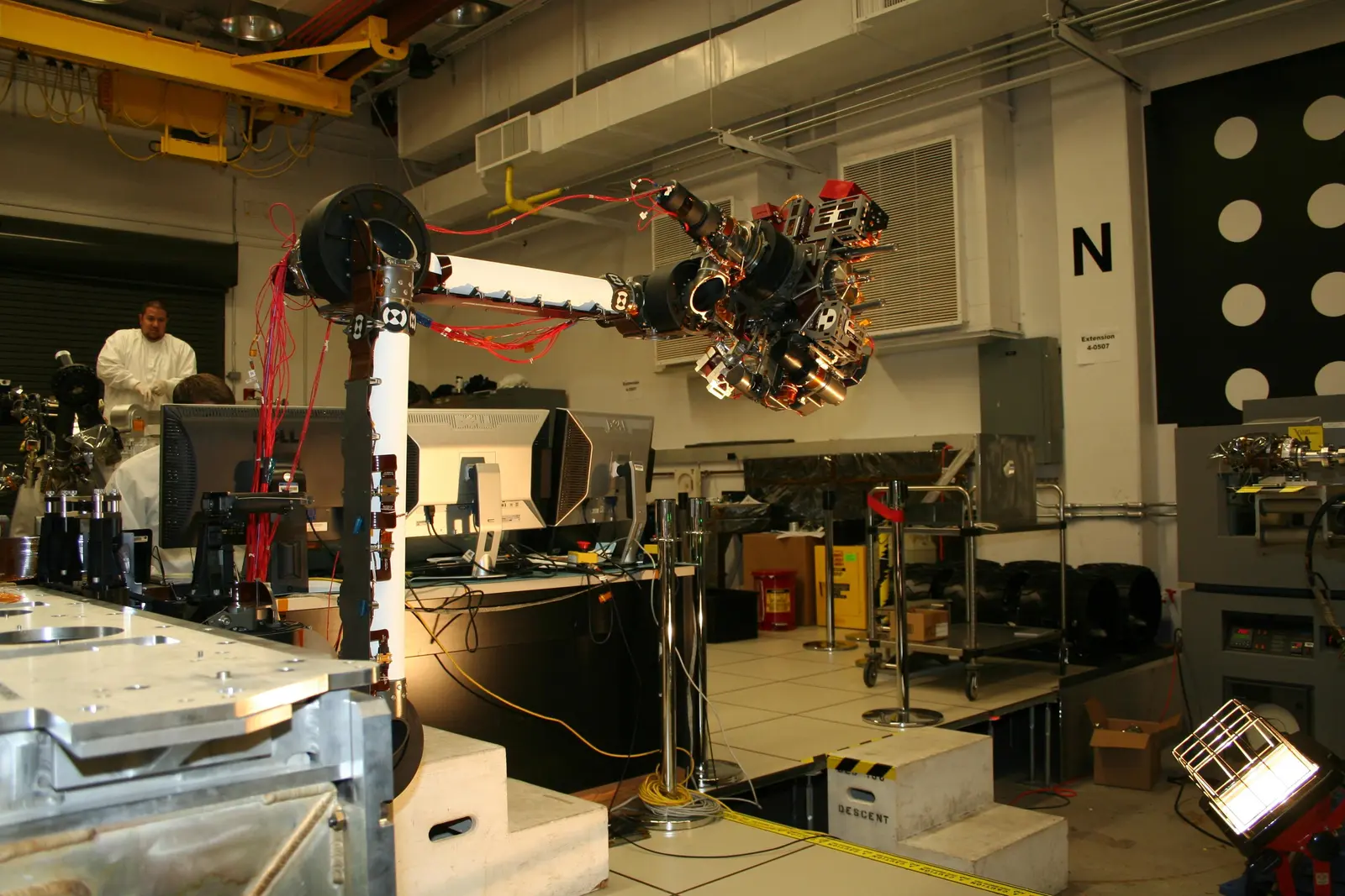 A complex robotic arm with exposed wiring and sensors is mounted on a stand in a laboratory setting, with a person in a white coat visible in the background. The photographer titled it “Curiosity Robotic Arm Bent at Nearly a 90-degree Angle”.