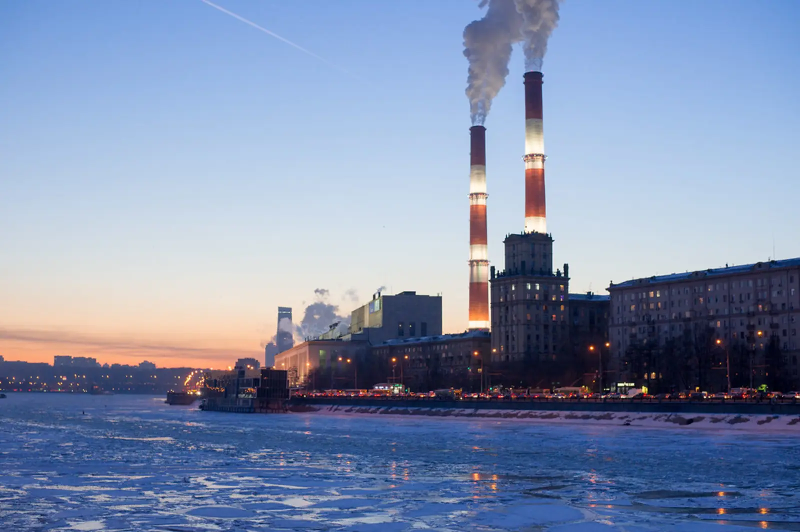 Smoke billows from two tall, striped chimneys of a power plant situated along a frozen riverbank under a twilight sky. The photographer titled it “Power plant №12 in winter”.