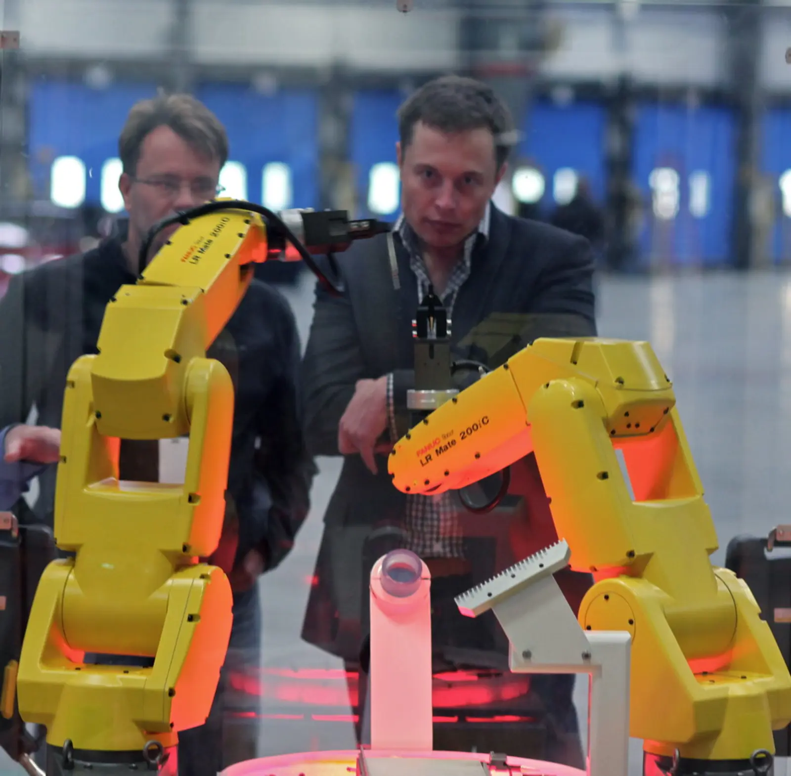 Two yellow articulated robotic arms are positioned in the foreground, while two men stand behind them observing the display, with reflections visible on the glass surface in front of the scene. The photographer titled it “FANUC Robot Assembly Demo for Elon at Tesla”.