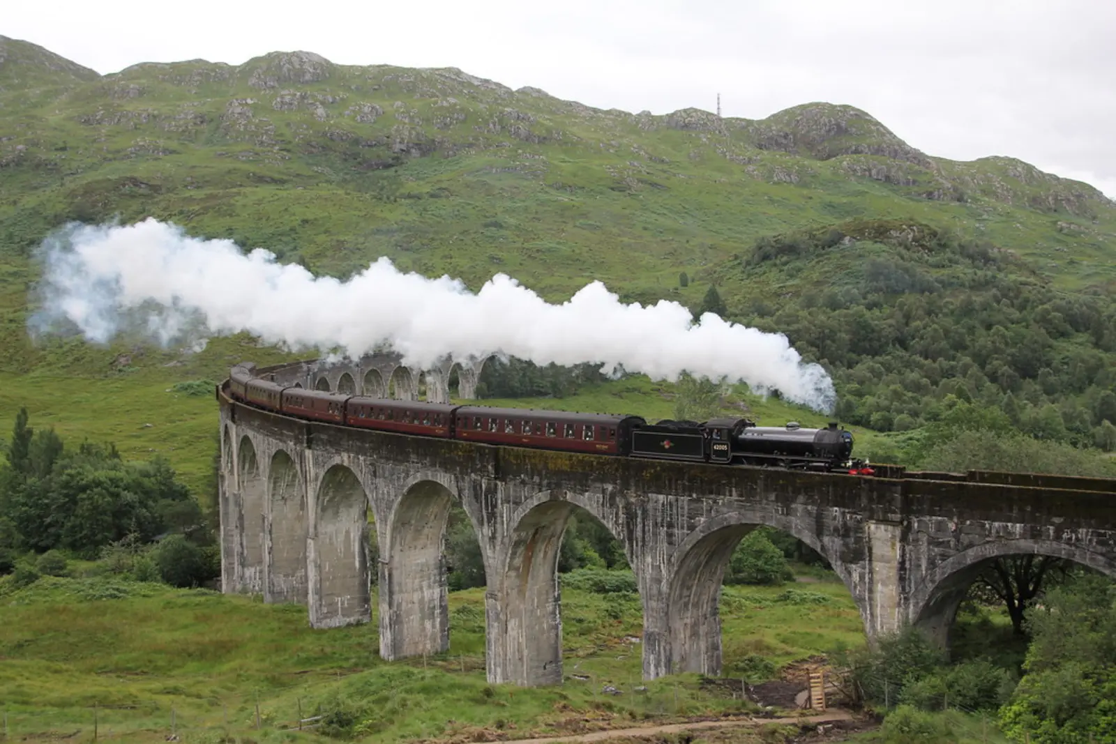 A black steam locomotive pulling red passenger carriages crosses a curved, multi-arched viaduct, emitting a large plume of white smoke against a backdrop of green hills.