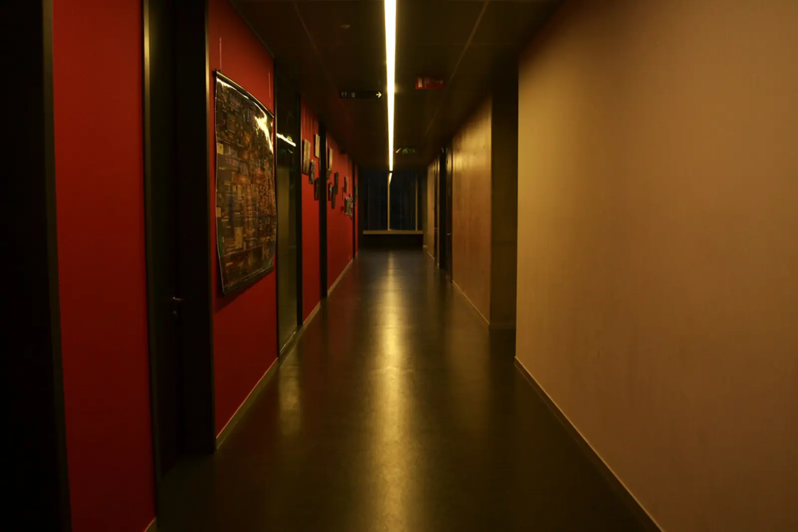 An empty hallway with a red wall on the left displaying posters and a beige wall on the right, illuminated by a long strip light on the ceiling. The photographer titled it “School”.