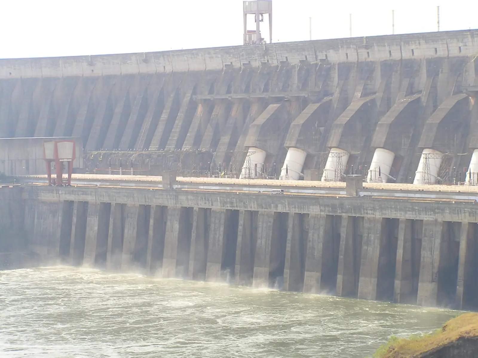 A large concrete dam with multiple spillways is shown releasing water into a turbulent river below.