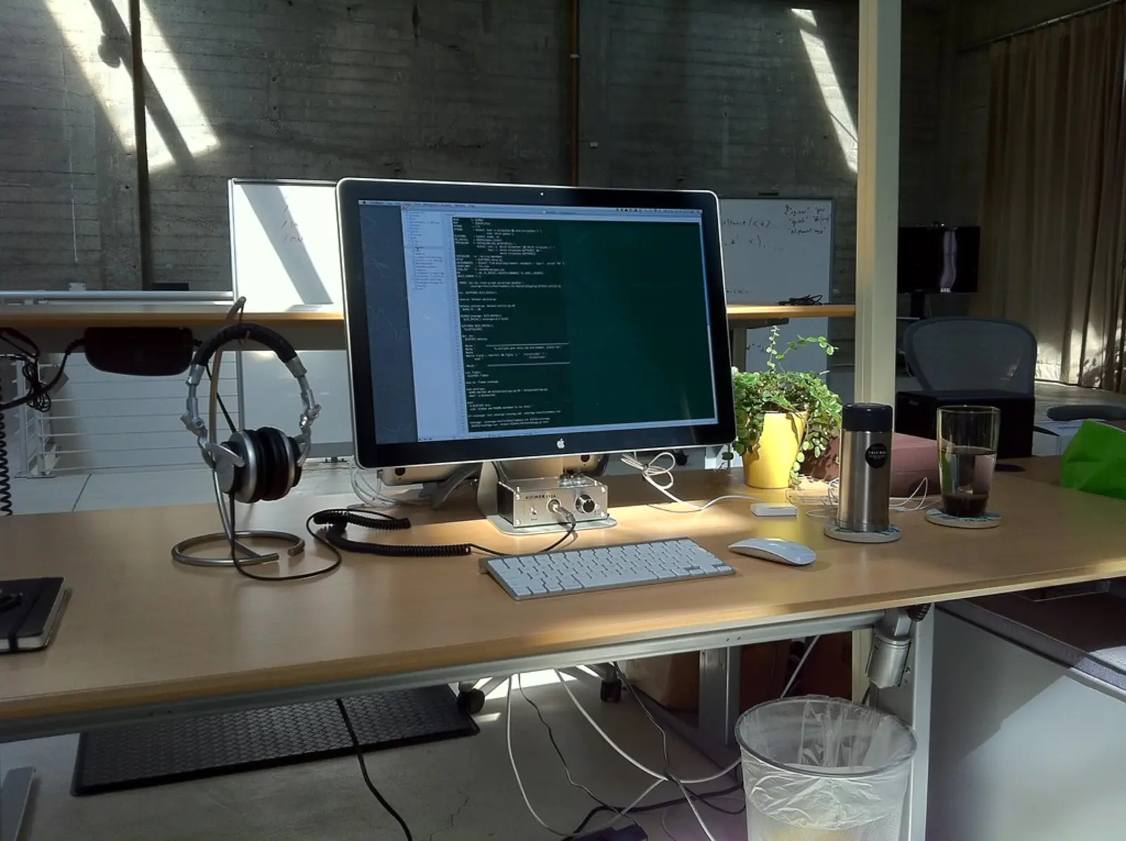 A computer monitor displaying lines of code on a dark screen sits on a wooden desk alongside a keyboard, mouse, headphones, and a cup of coffee. The photographer titled it “My Desk @ SimpleGeo SF”.