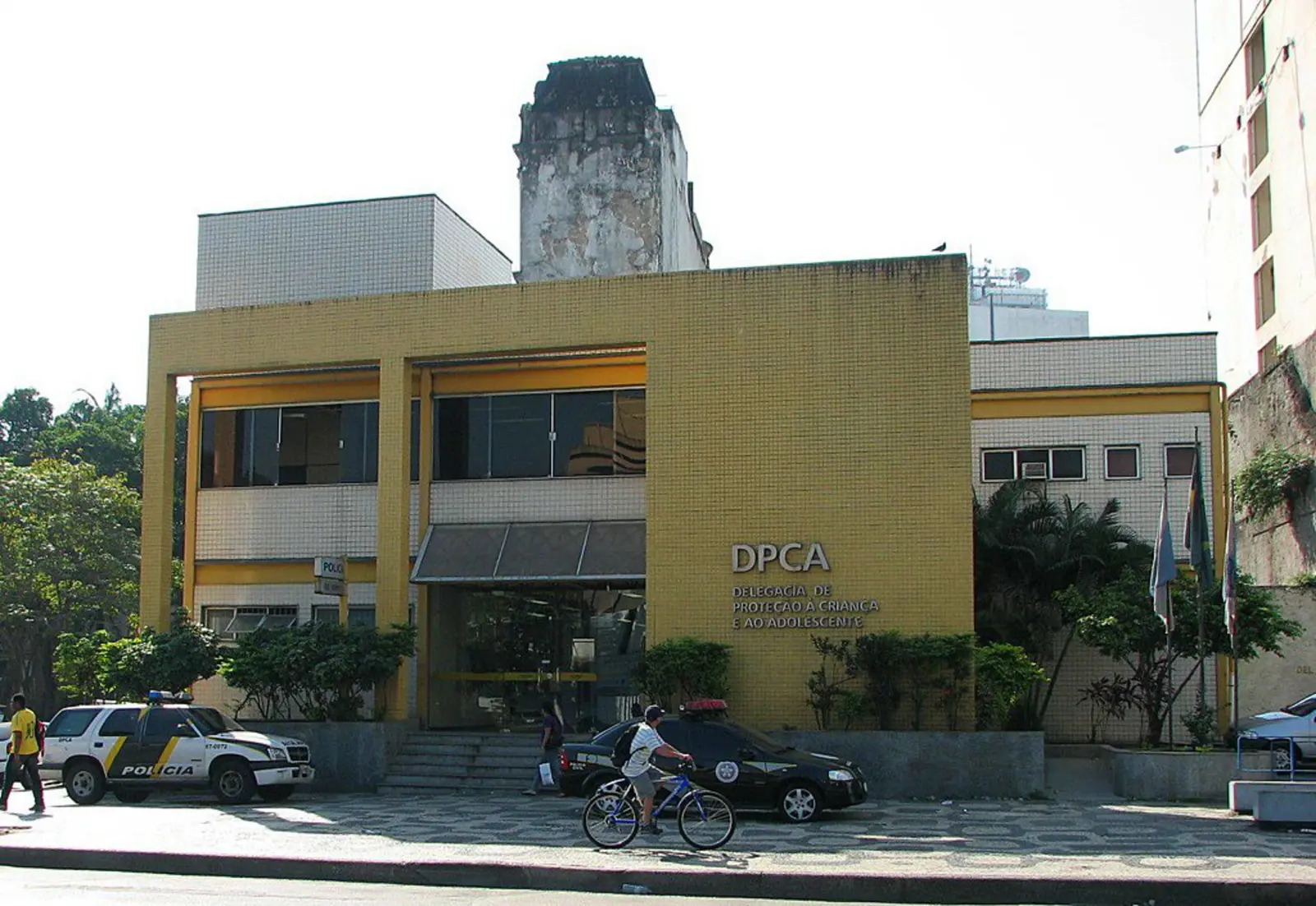 A cyclist rides past a yellow police station building marked 'DPCA', with a police vehicle parked outside. The photographer titled it “Delegacia de Polícia - Polícia Civil - Rio de Janeiro”.