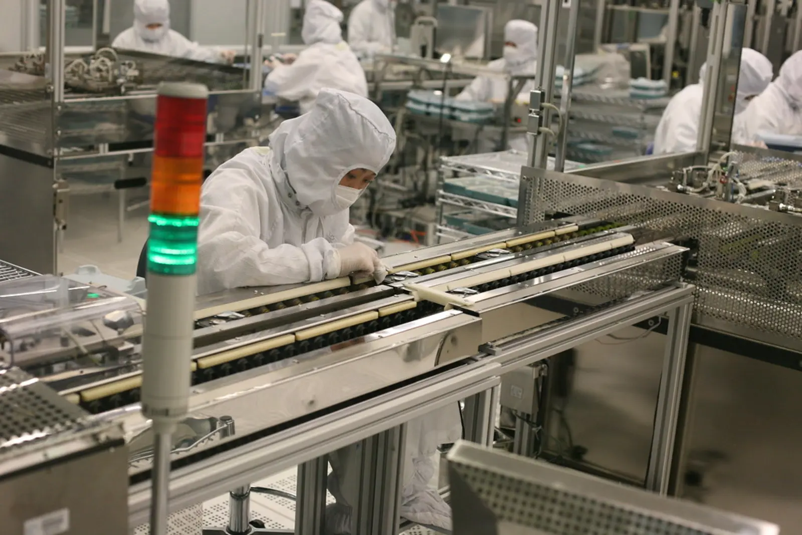 A worker in a full protective cleanroom suit operates machinery along a conveyor belt in a manufacturing facility, with a multi-colored signal light in the foreground. The photographer titled it “Seagate's clean room”.
